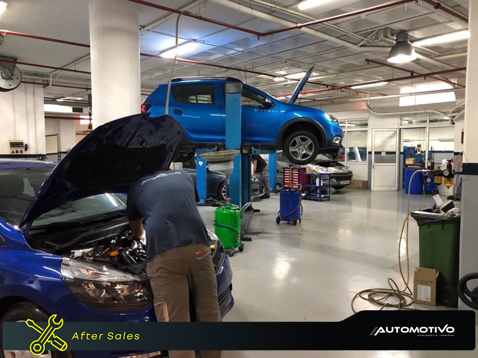 a man wearing a blue t-shirt and brown pants fixing a blue car while behind him there is another car to fix