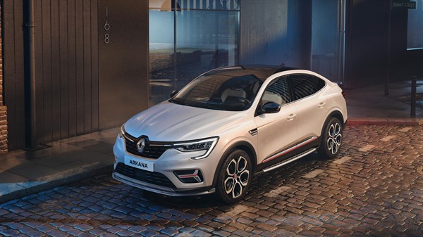 a man is sitting in the edge of a road. Infront of him he has the renault Arkana full hybrid car. Behind him you can see the sea.