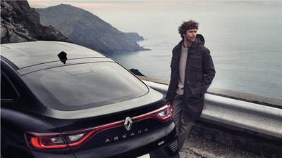 a man is sitting in the edge of a road. Infront of him he has the renault Arkana full hybrid car. Behind him you can see the sea.